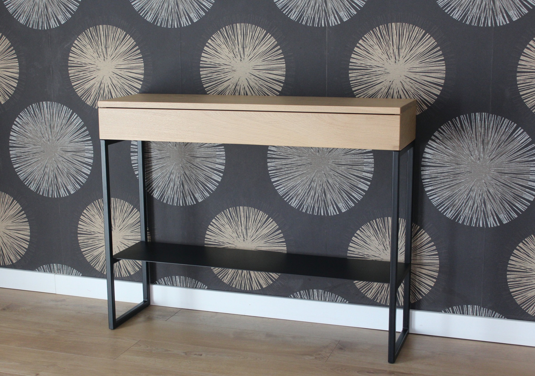 Konstrukcja konsoli z jasnego drewna z szufladą i czarnymi metalowymi nogami na tle tapety w geometryczne wzory.