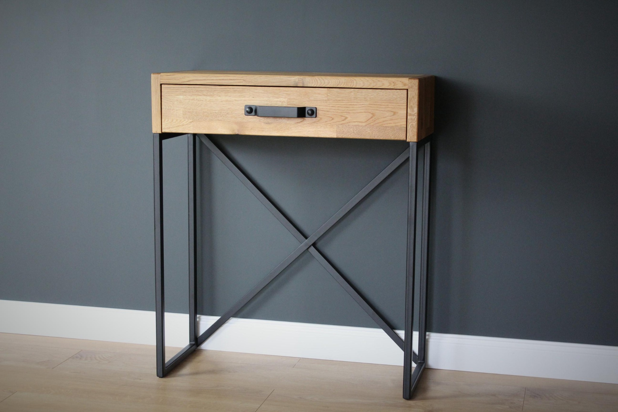 Drewniana szafka nocna z pojedynczą szufladą i metalowym stelażem w kształcie litery X, na tle szarej ściany i jasnej podłogi.