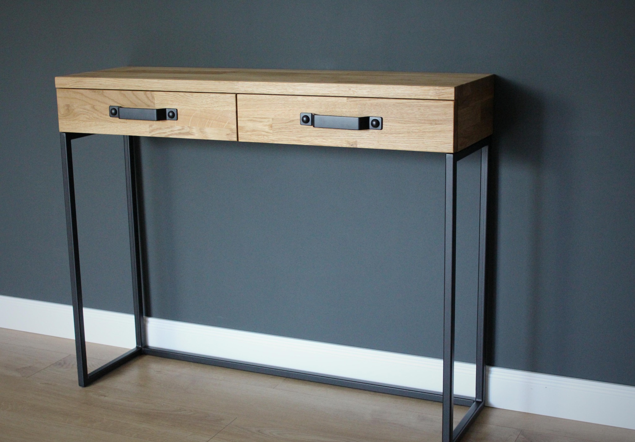 Konsola z dwoma szufladami z jasnego drewna na metalowym, czarnym stelażu, minimalistyczny design na tle szarej ściany.