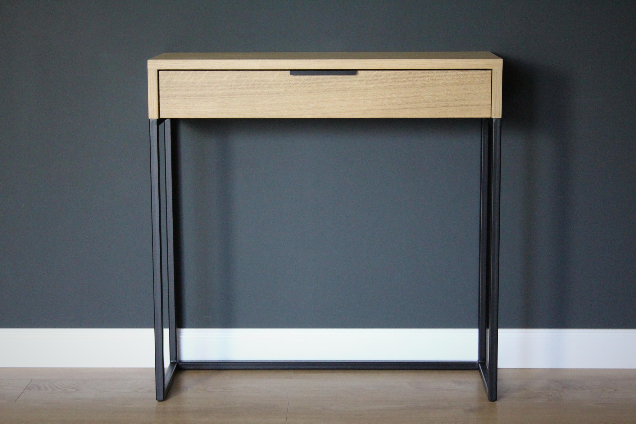 Konsola z jasnego drewna z szufladą i czarnymi metalowymi nogami na tle szarej ściany.