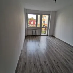 Pusty pokój po remoncie z szarą podłogą imitującą drewno, białymi ścianami i grzejnikiem pod oknem z widokiem na budynki.