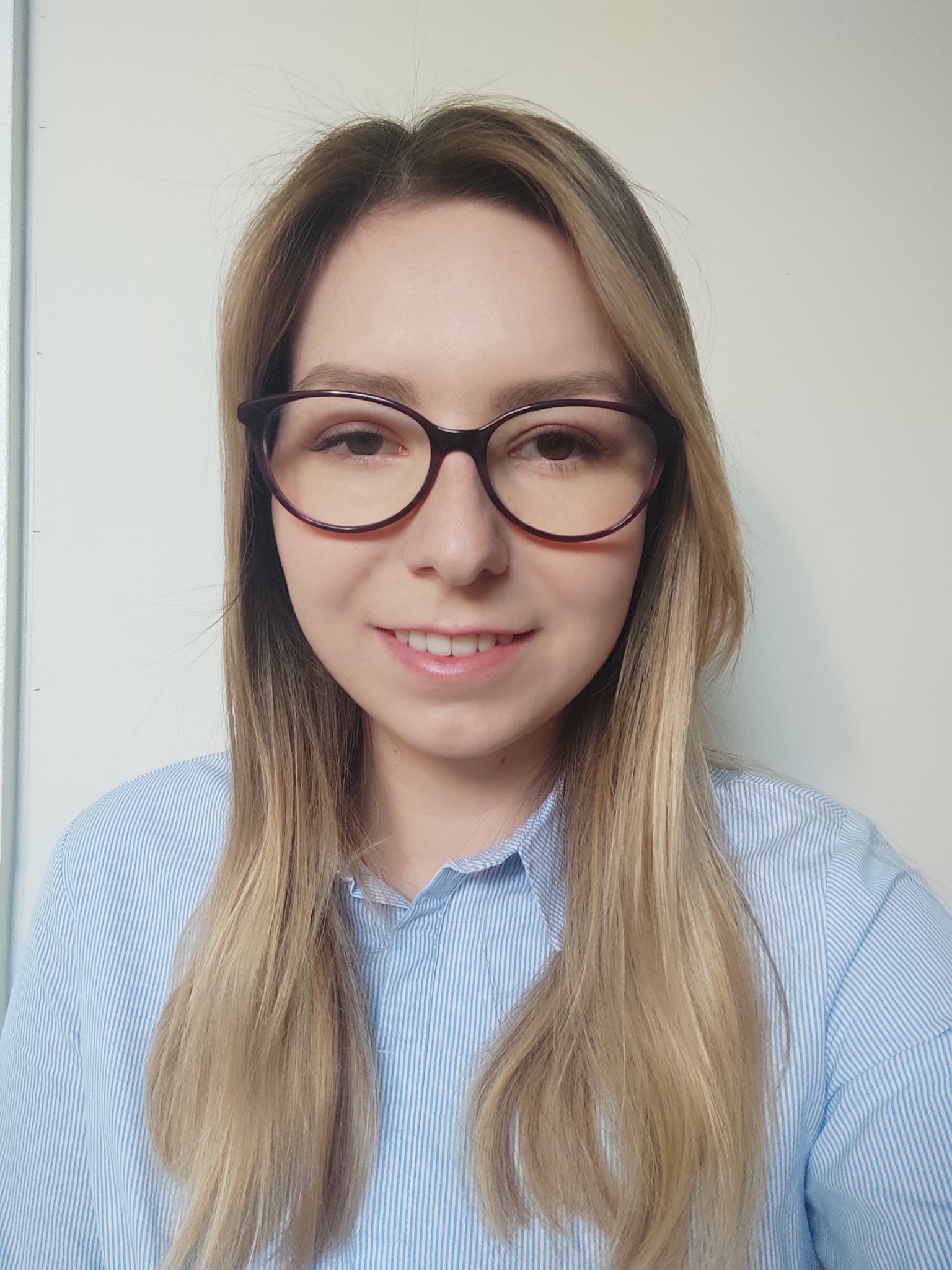 Portret uśmiechniętej blondynki w okularach w bordowej oprawce i błękitnej koszuli w paski, na jasnym tle.