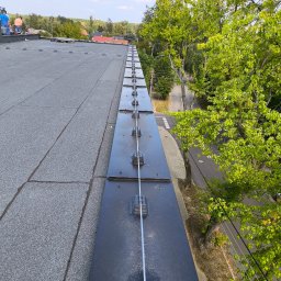 Elektryk Przemysław Nurkiewicz - Dach budynku z zamontowaną instalacją odgromową, widok z góry, z zielenią drzew w tle. Elementy instalacji równo rozmieszczone wzdłuż krawędzi dachu.