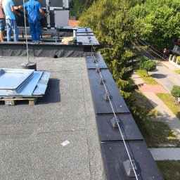 Elektryk Przemysław Nurkiewicz - Montaż instalacji odgromowej na dachu budynku. Widoczne uchwyty, przewody i ekipa montażystów przy pracy. W tle drzewa i ulica.