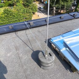 Elektryk Przemysław Nurkiewicz - Instalacja odgromowa na dachu budynku z widocznym uziomem i przewodami odprowadzającymi. W tle zabudowa mieszkalna i zieleń.