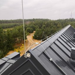 Elektryk Przemysław Nurkiewicz - Szara blacha na dachu z zamontowaną instalacją odgromową, widok z góry na las w tle. Metalowe elementy instalacji są przymocowane do dachu.