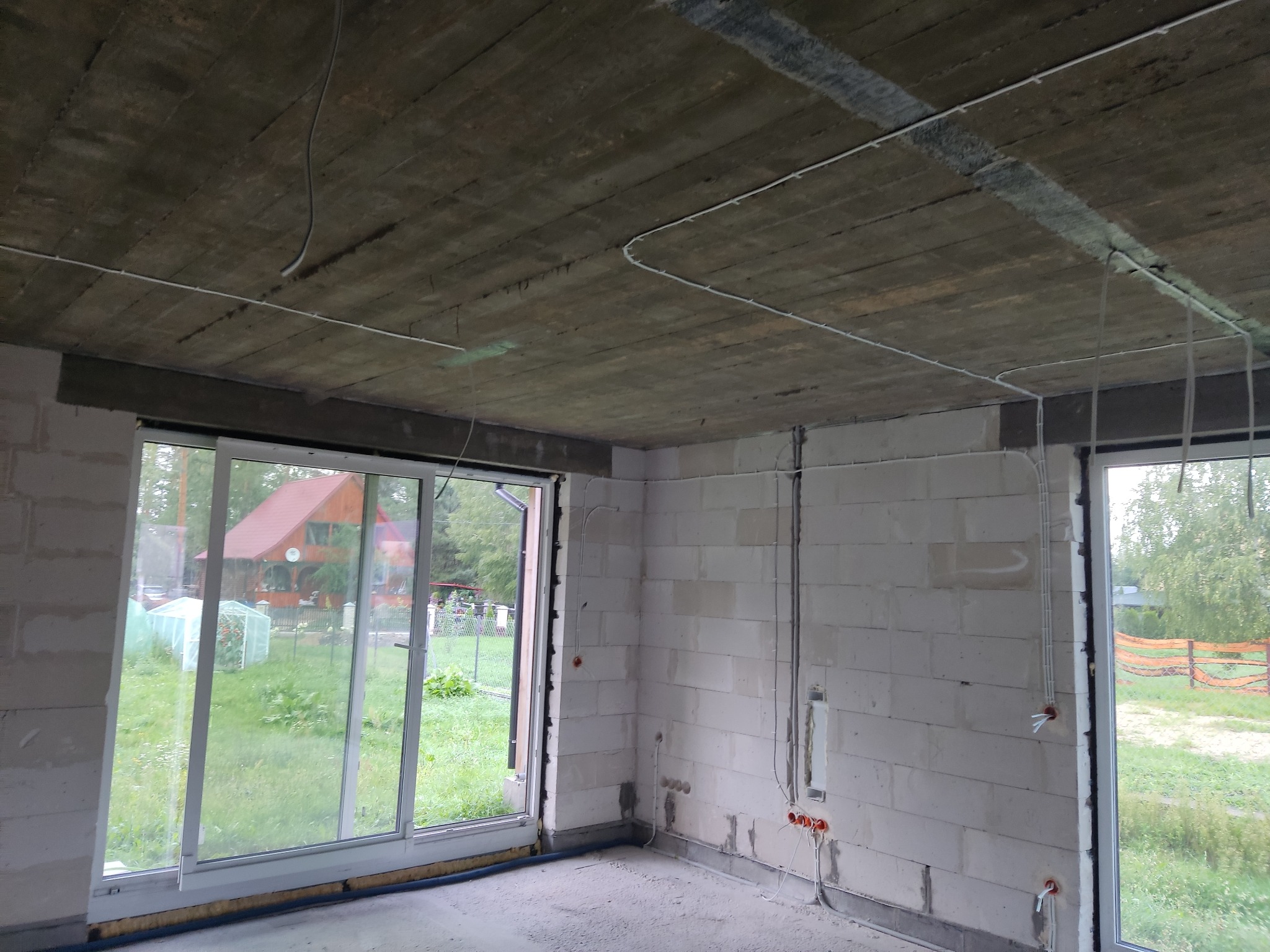 Surowy stan wnętrza z widocznymi przewodami elektrycznymi na suficie i ścianach, okna z widokiem na zieleń, ściany z betonu komórkowego.