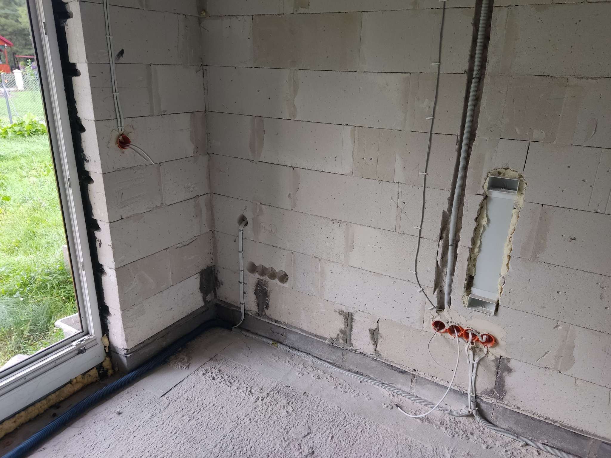 Surowy stan: instalacja elektryczna w ścianach z betonu komórkowego. Widoczne kable, puszki i peszle przed tynkowaniem. Okno z widokiem na zieleń.