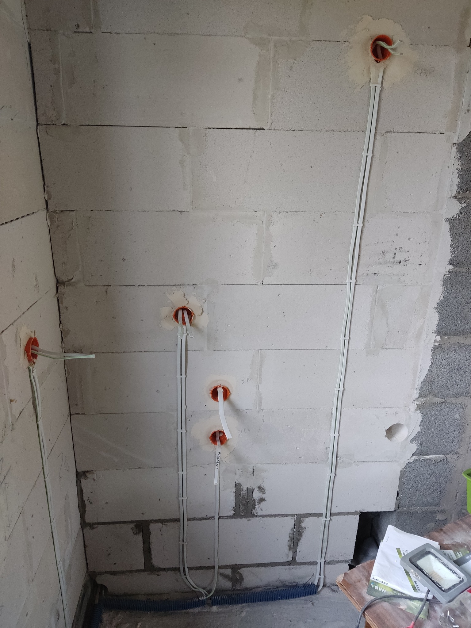 Surowa ściana z betonu komórkowego z widocznymi, świeżo poprowadzonymi przewodami w peszlach, zakończonymi pomarańczowymi puszkami instalacyjnymi.