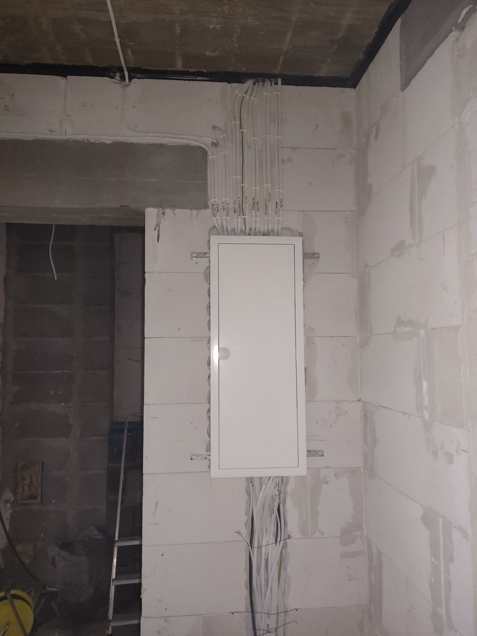 Biała skrzynka elektryczna z licznymi przewodami w surowym wnętrzu, widoczne przewody w ścianie i suficie. Na ścianie widoczne napisy na przewodach.