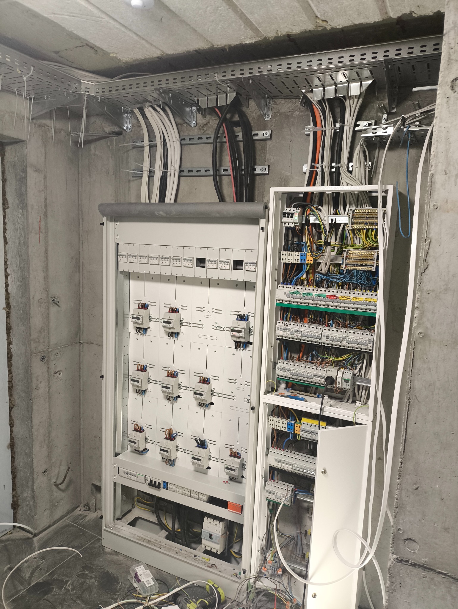Rozdzielnia elektryczna z licznikami, kable prowadzone w korytach na surowym suficie. Widoczne zabezpieczenia i okablowanie. Stan surowy, betonowe ściany.