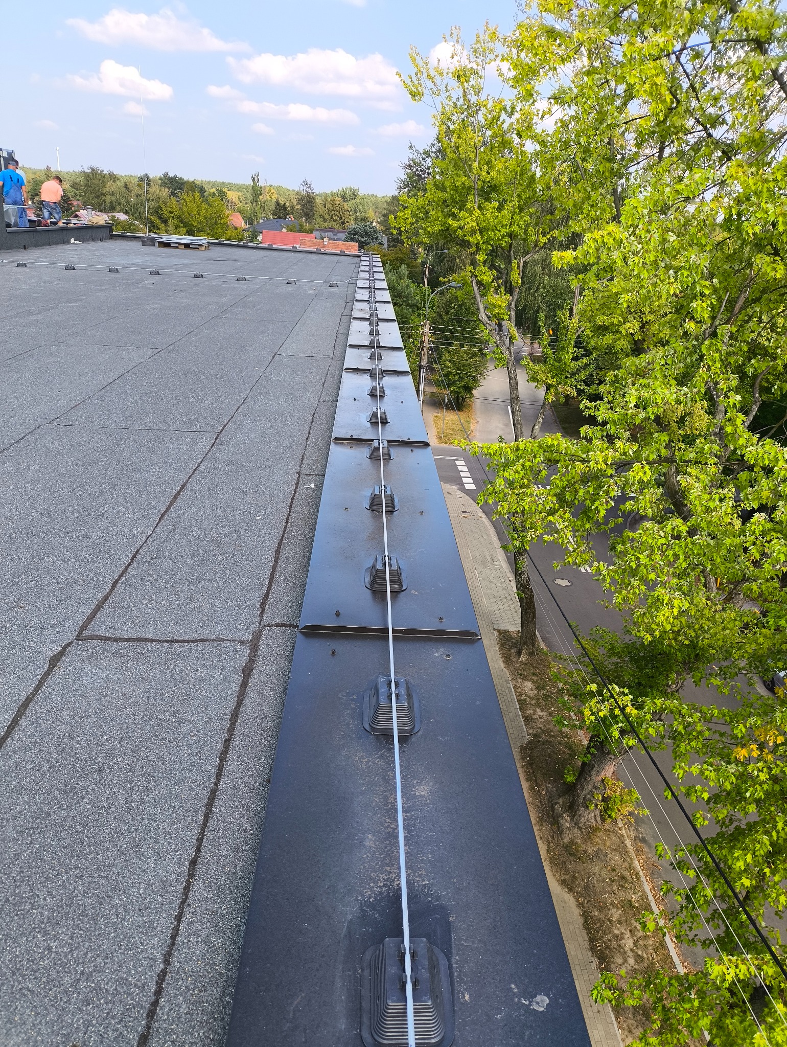Dach budynku z zamontowaną instalacją odgromową, widok z góry, z zielenią drzew w tle. Elementy instalacji równo rozmieszczone wzdłuż krawędzi dachu.