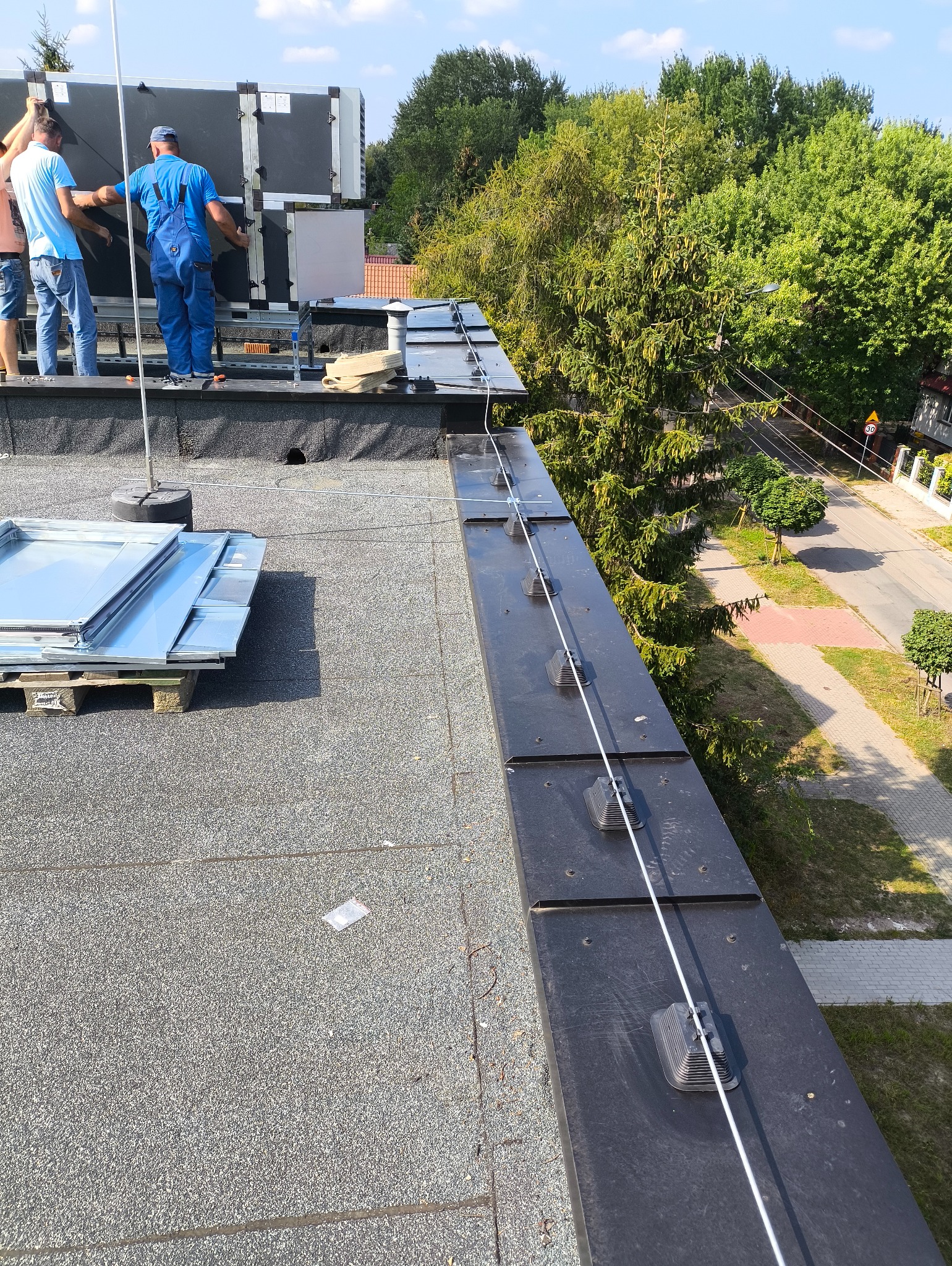 Montaż instalacji odgromowej na dachu budynku. Widoczne uchwyty, przewody i ekipa montażystów przy pracy. W tle drzewa i ulica.