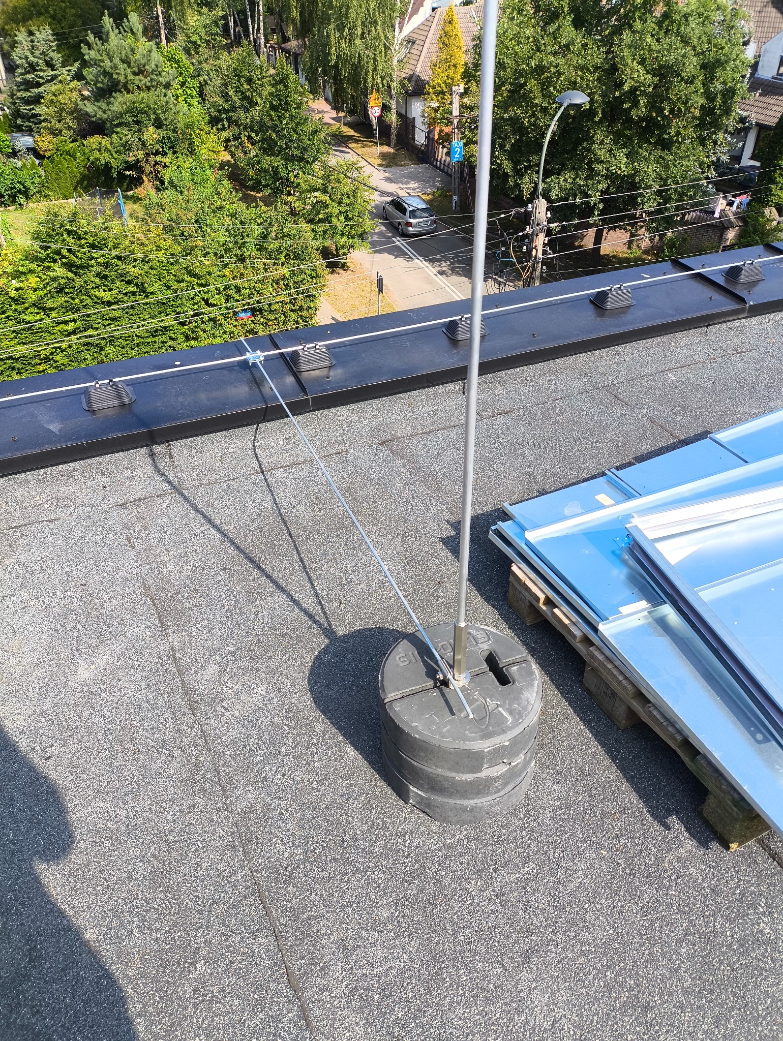 Instalacja odgromowa na dachu budynku z widocznym uziomem i przewodami odprowadzającymi. W tle zabudowa mieszkalna i zieleń.