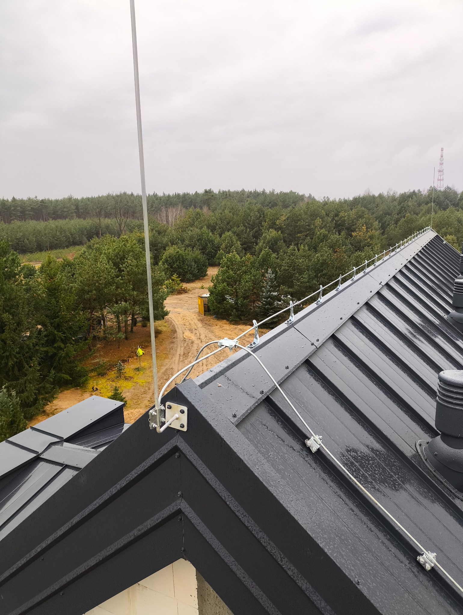 Szara blacha na dachu z zamontowaną instalacją odgromową, widok z góry na las w tle. Metalowe elementy instalacji są przymocowane do dachu.