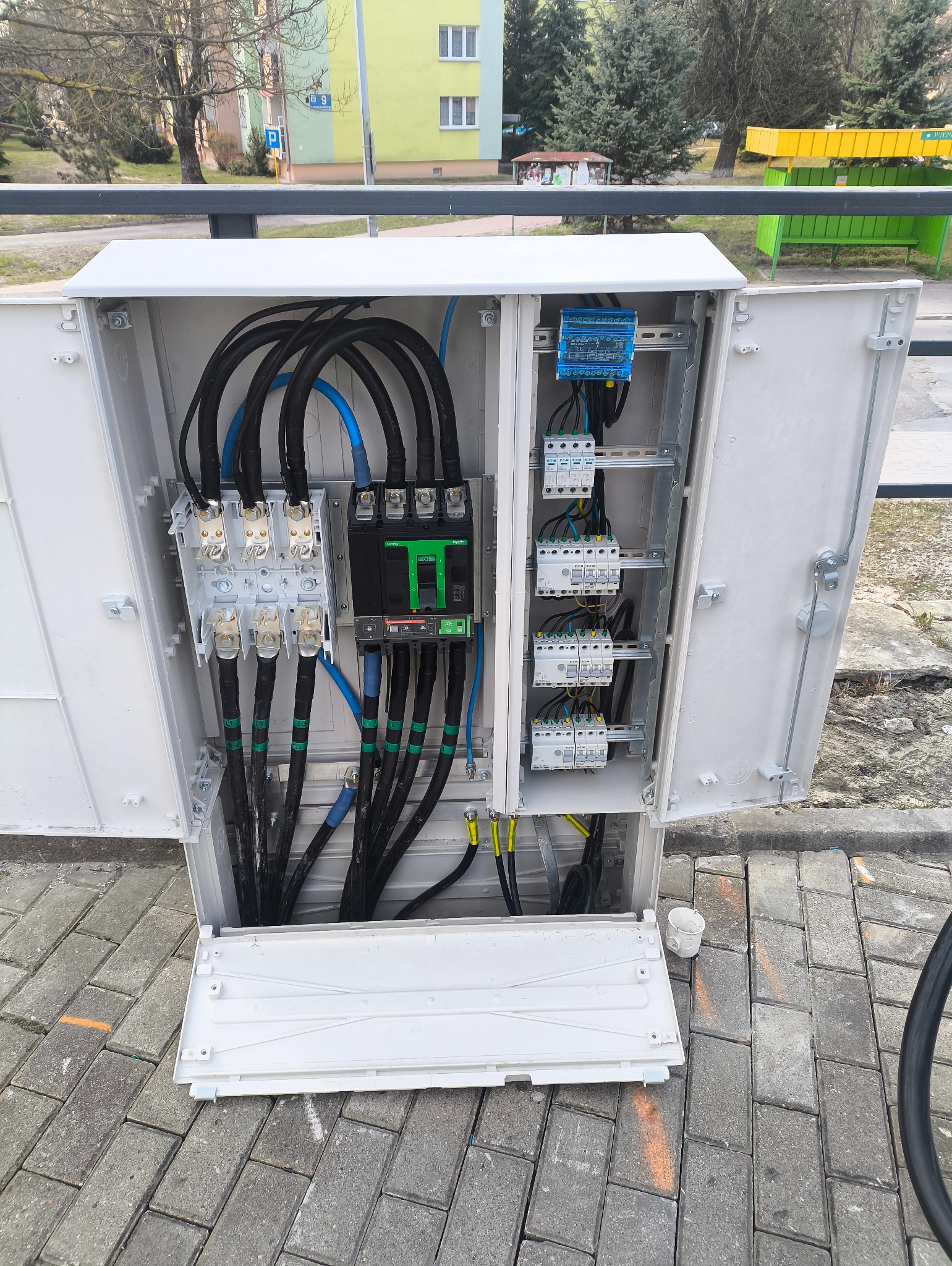 Otwarta szafa elektryczna na zewnątrz z widocznymi kablami, bezpiecznikami i elementami instalacji. Widoczne kable w izolacji czarnej, niebieskiej, zielonej i żółtej.