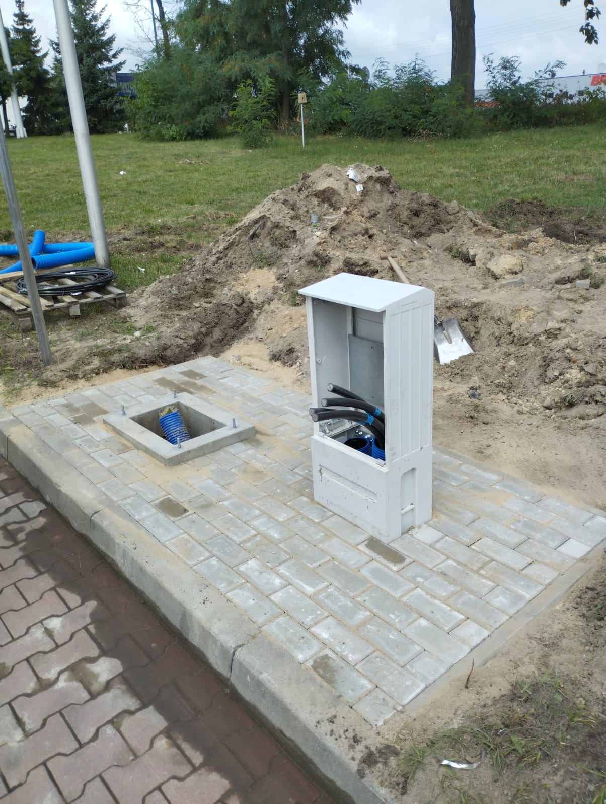 Szafka elektryczna z okablowaniem w fundamencie z kostki brukowej na zewnątrz, w tle widoczna ziemia i zieleń.