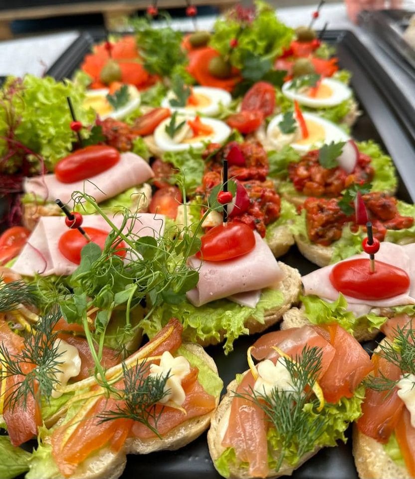Różnorodne kanapki na czarnym półmisku: z łososiem, szynką, jajkiem, pastą warzywną, udekorowane sałatą, pomidorkami cherry i świeżymi ziołami, spięte ozdobnymi wykałaczkami.