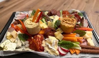 Dekoracyjna taca z różnorodnymi przekąskami: sery, wędliny, warzywa (marchew, rzodkiewka, seler naciowy), hummus w szklanych naczyniach, ułożone na białym papierze na czarnej tacy.