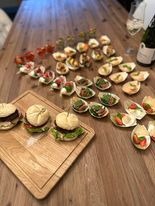 Drewniany stół z różnorodnymi, apetycznie wyglądającymi przekąskami: mini burgery na desce, kanapeczki w łódeczkach i szklanki z napojami w tle.