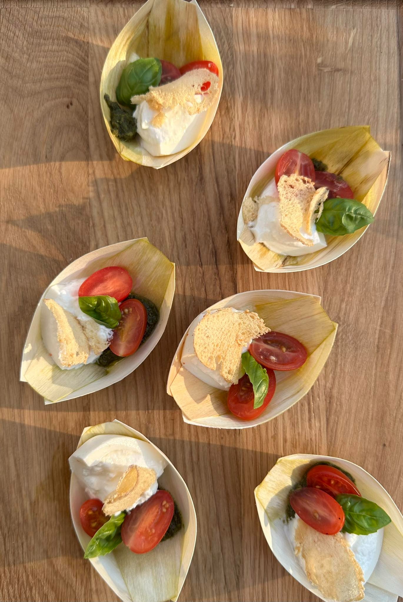 Elegancko podane przystawki: mini porcje caprese z mozzarellą, bazylią, pomidorkami cherry i grzanką, serwowane w naturalnych łódeczkach z liści palmowych, na drewnianym stole w ciepłym świetle.