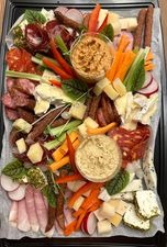 Bogato zdobiona taca cateringowa z różnorodnymi przekąskami: wędliny, sery, warzywa (marchew, seler naciowy, rzodkiewki), dipy w szklanych naczyniach, całość ułożona na papierze pergaminowym.