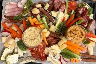 Bogato zaaranżowana deska serów, wędlin i warzyw, z dwoma miseczkami dipów, ułożona na jasnym papierze na czarnej tacy, widok z góry.