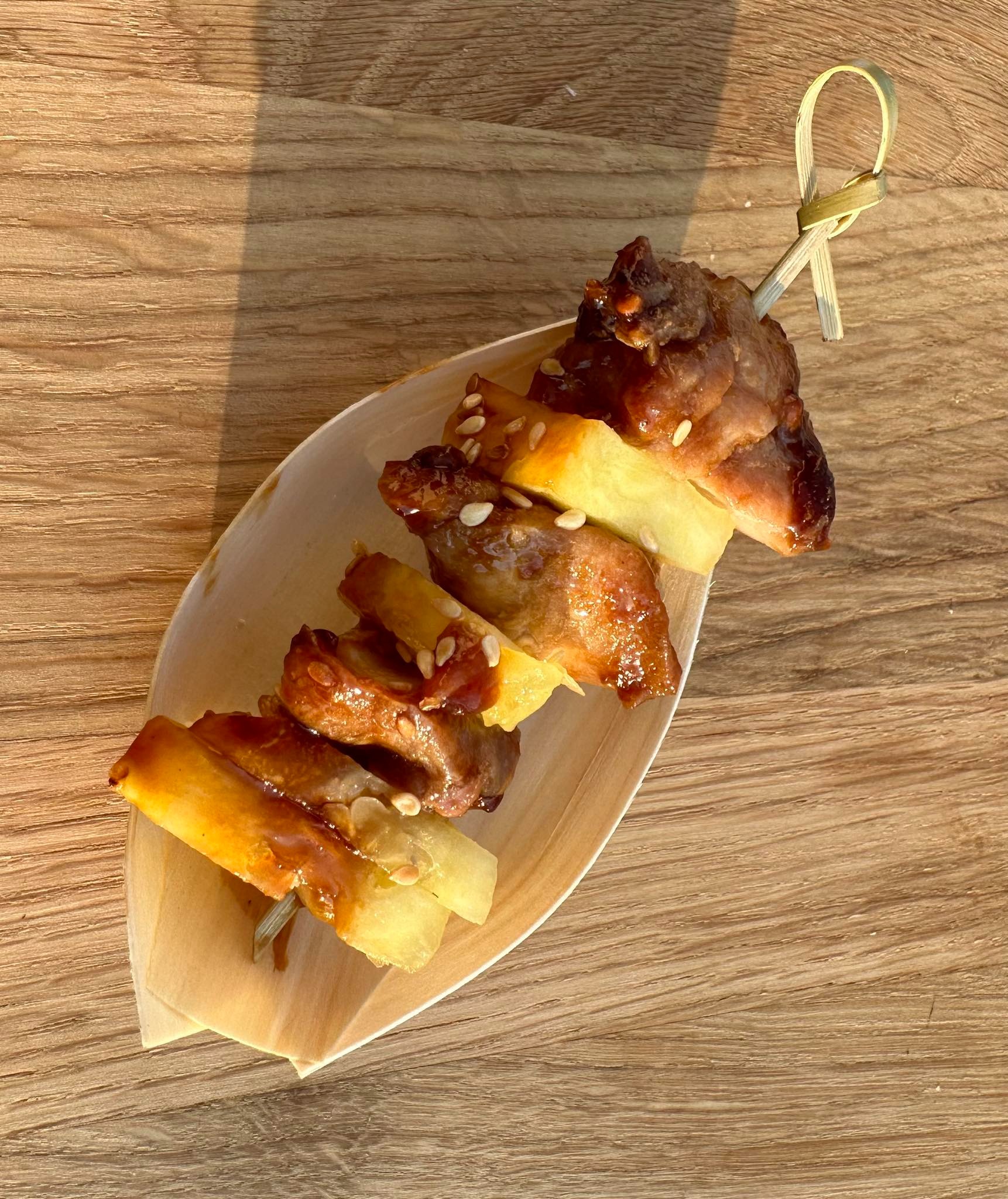 Miniaturowy szaszłyk z glazurowanego mięsa i ananasa, posypany sezamem, podany na łódce z jasnego drewna na drewnianym stole.