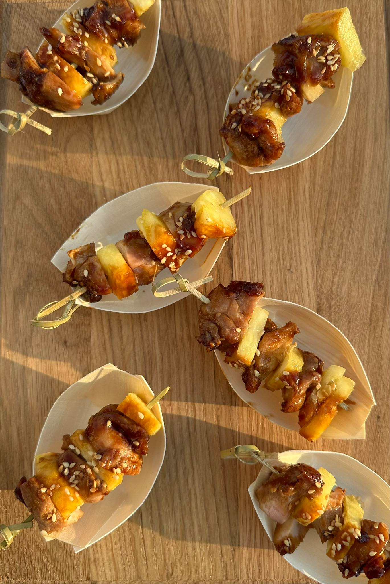 Mini szaszłyki z ananasem i mięsem w glazurze, posypane sezamem, podane na małych, ekologicznych łódeczkach z drewna na drewnianym stole.