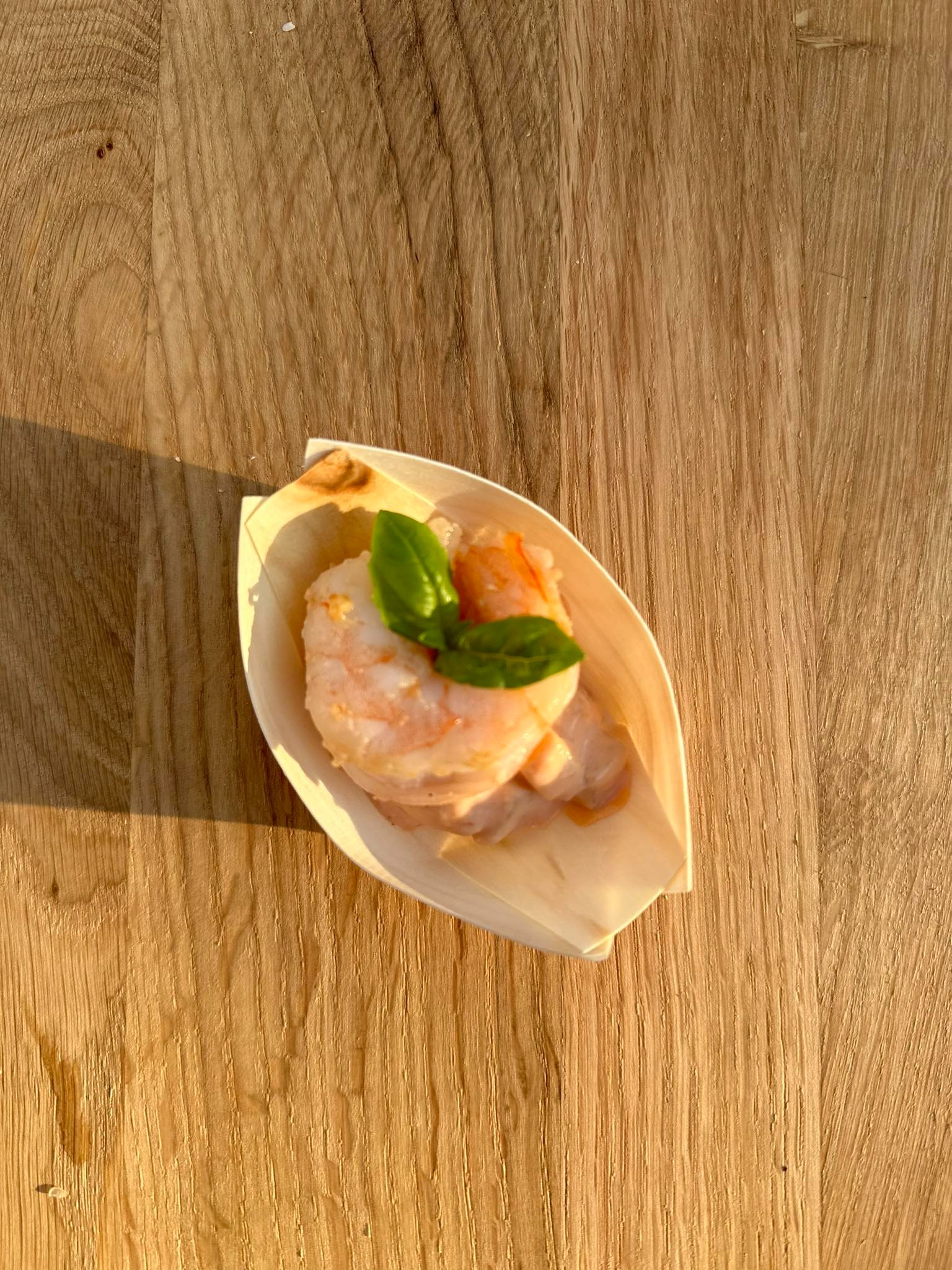 Pojedyncza porcja krewetek w sosie, udekorowana listkiem bazylii, serwowana w małej, drewnianej łódeczce na tle drewnianego blatu w ciepłym oświetleniu.