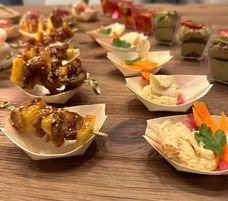 Elegancko podane przekąski cateringowe w małych, drewnianych miseczkach na stole, widoczne szaszłyki z ananasem i mięsem, pasta z warzywami i inne małe dania.