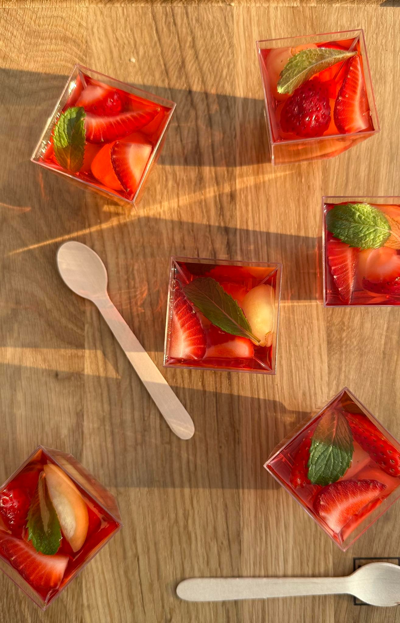 Deser w przezroczystych kubeczkach z galaretką truskawkową, kawałkami truskawek, melona i listkami mięty, ułożony na drewnianym stole z dwiema drewnianymi łyżeczkami, oświetlony ciepłym światłem...