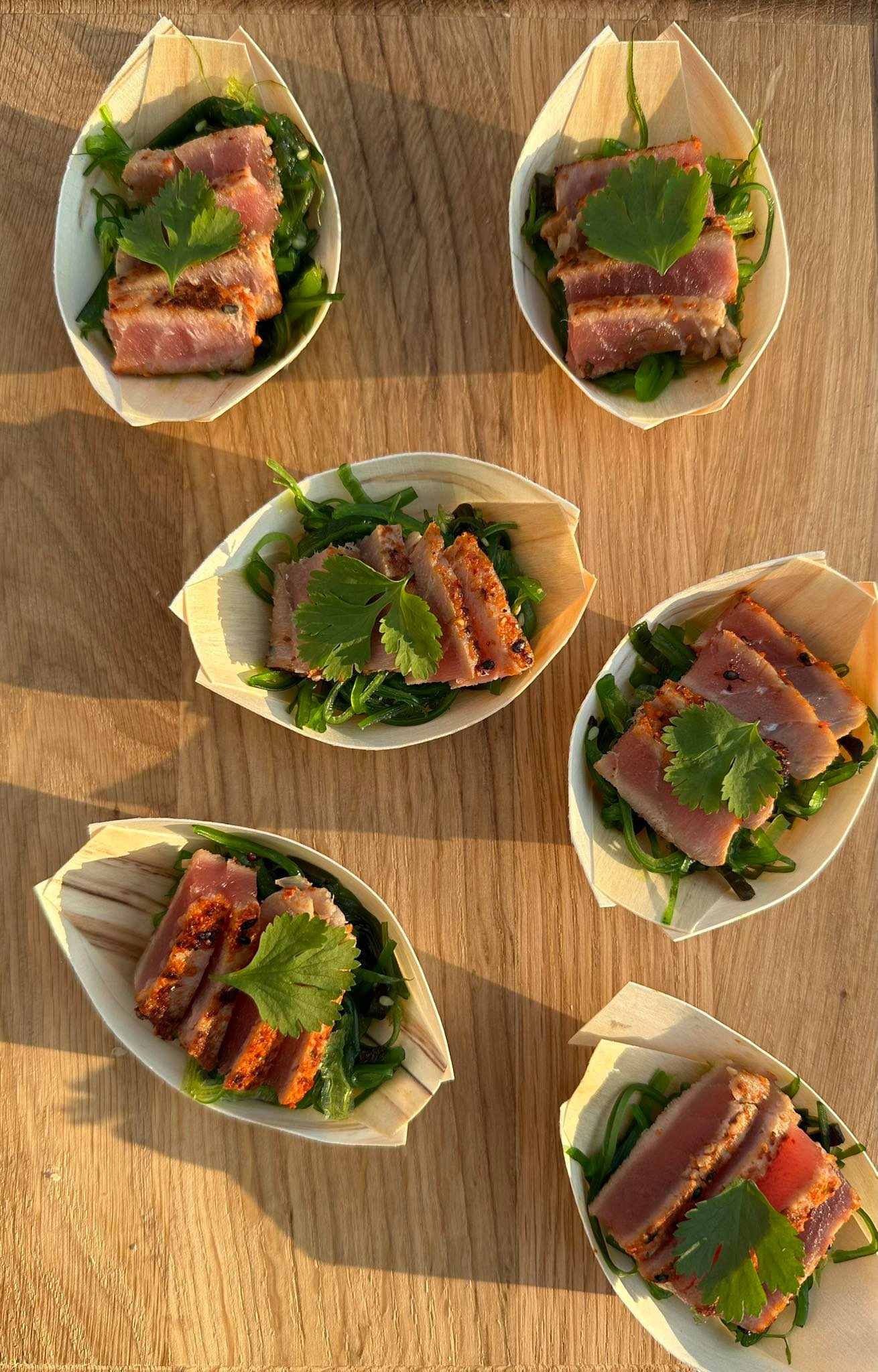 Apetizing porcje grillowanego tuńczyka na sałatce z wodorostów, podane w łódeczkach z jasnego drewna, ułożone na drewnianym stole w ciepłym świetle.