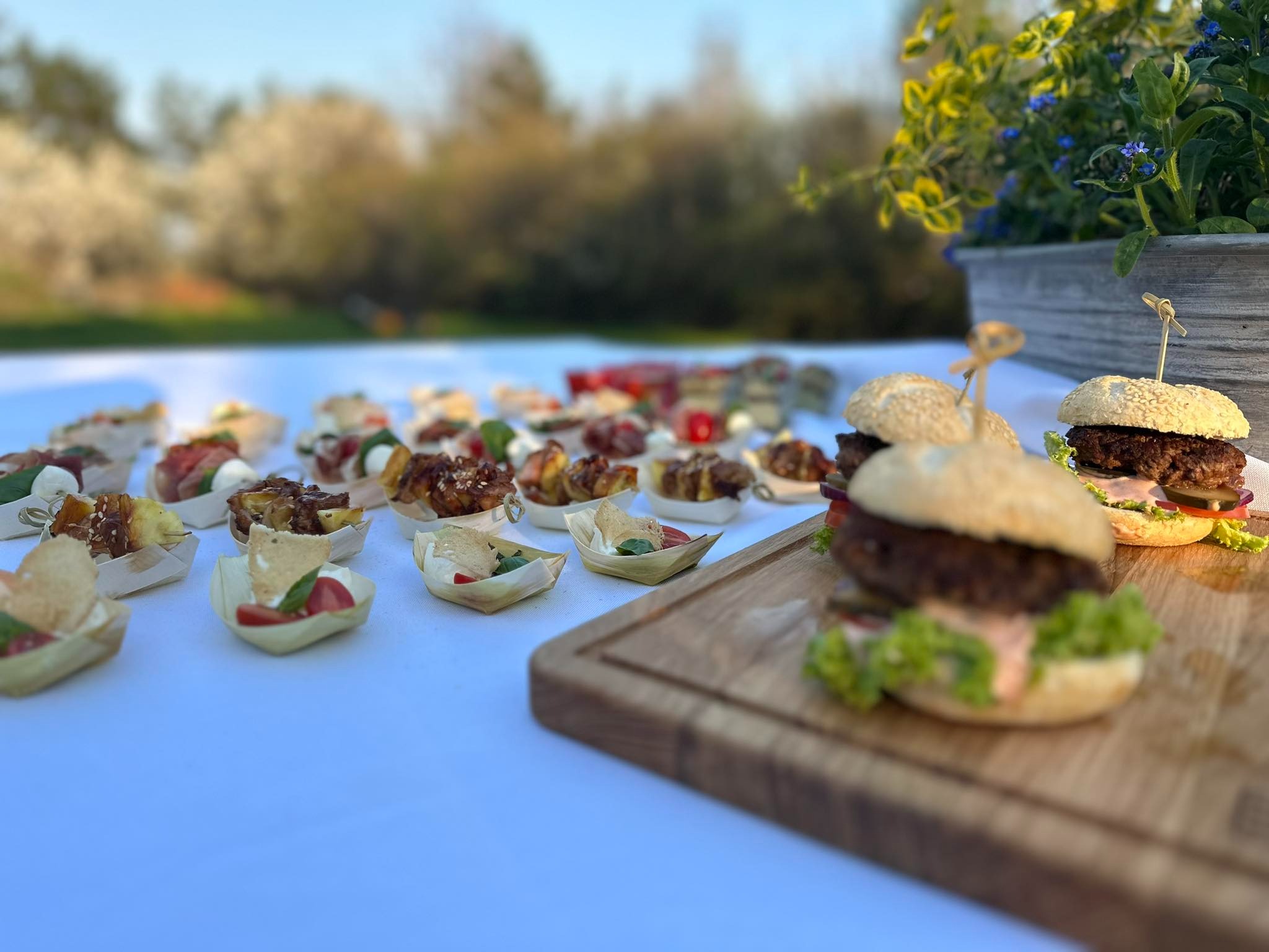 Elegancko podane mini burgery i przekąski w drewnianych łódeczkach na białym obrusie, z kwitnącymi drzewami w tle, prezentacja plenerowa.