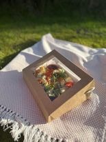 Kwadratowe pudełko z okienkiem wypełnione sałatką warzywną na białym kocu piknikowym, rozłożonym na trawie w słoneczny dzień.