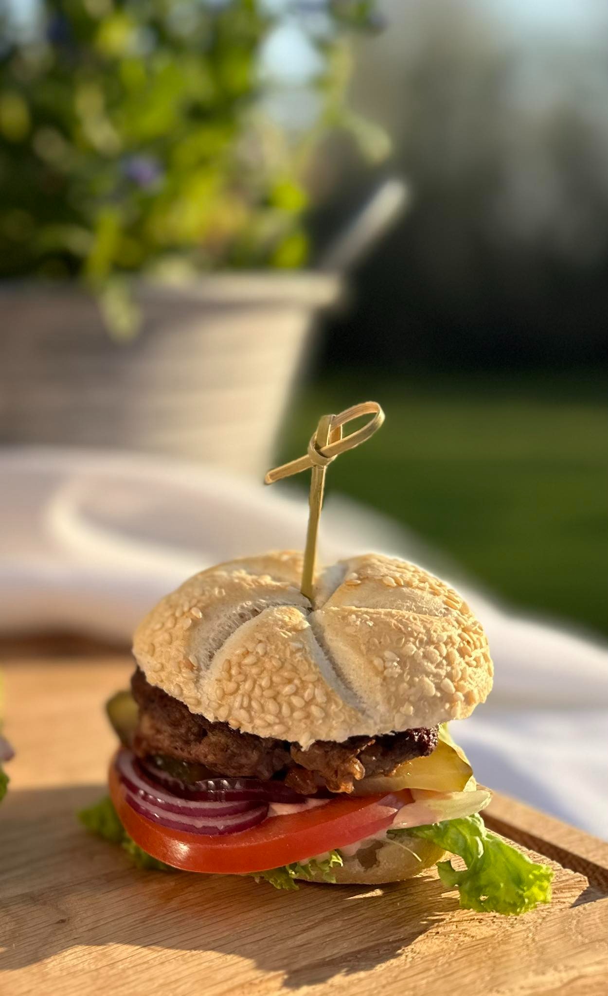 Mini burger z wołowiną, pomidorem, czerwoną cebulą, sałatą i ogórkiem kiszonym, spięty dekoracyjną wykałaczką, serwowany na drewnianej desce na tle ogrodu.