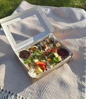 Otwarty kartonowy pojemnik z przekąskami na piknik, widoczne sery, owoce, warzywa i zielenina, ułożony na białym kocu w trawie, delikatne popołudniowe światło.