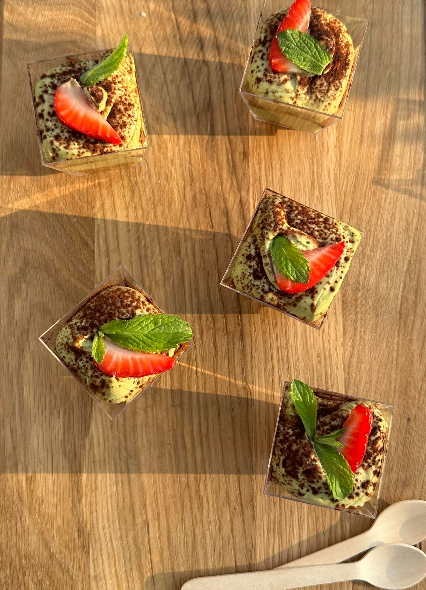 Elegancko podane desery w przezroczystych pojemnikach, z kremem pistacjowym, posypką kakaową, truskawką i miętą, ułożone na drewnianym blacie w ciepłym świetle.