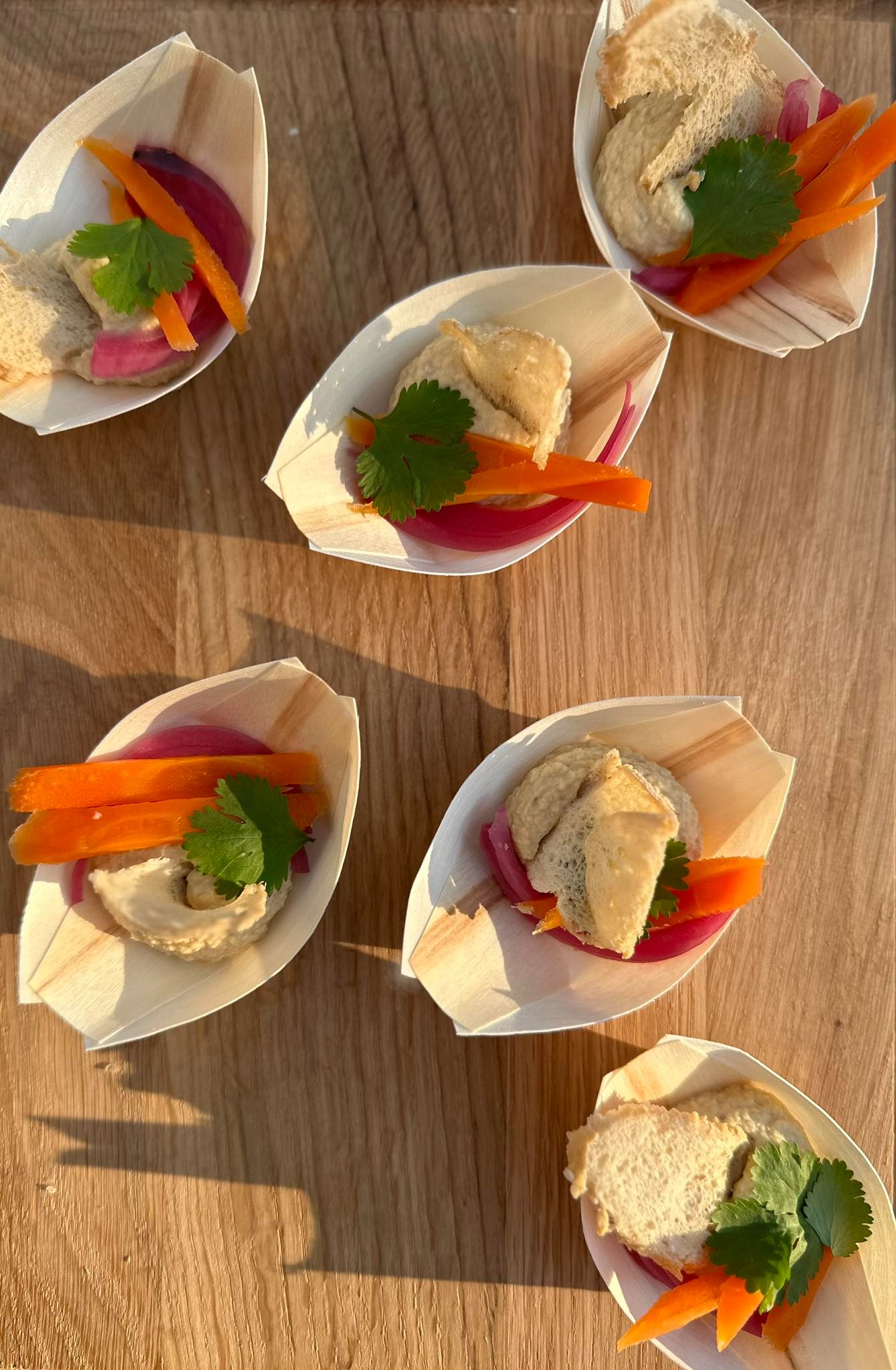 Degustacyjne przekąski: hummus z warzywami (marchewka, czerwona cebula) i pieczywem, podane w ekologicznych, drewnianych łódeczkach, udekorowane świeżą kolendrą, na drewnianym blacie w ciepłym...