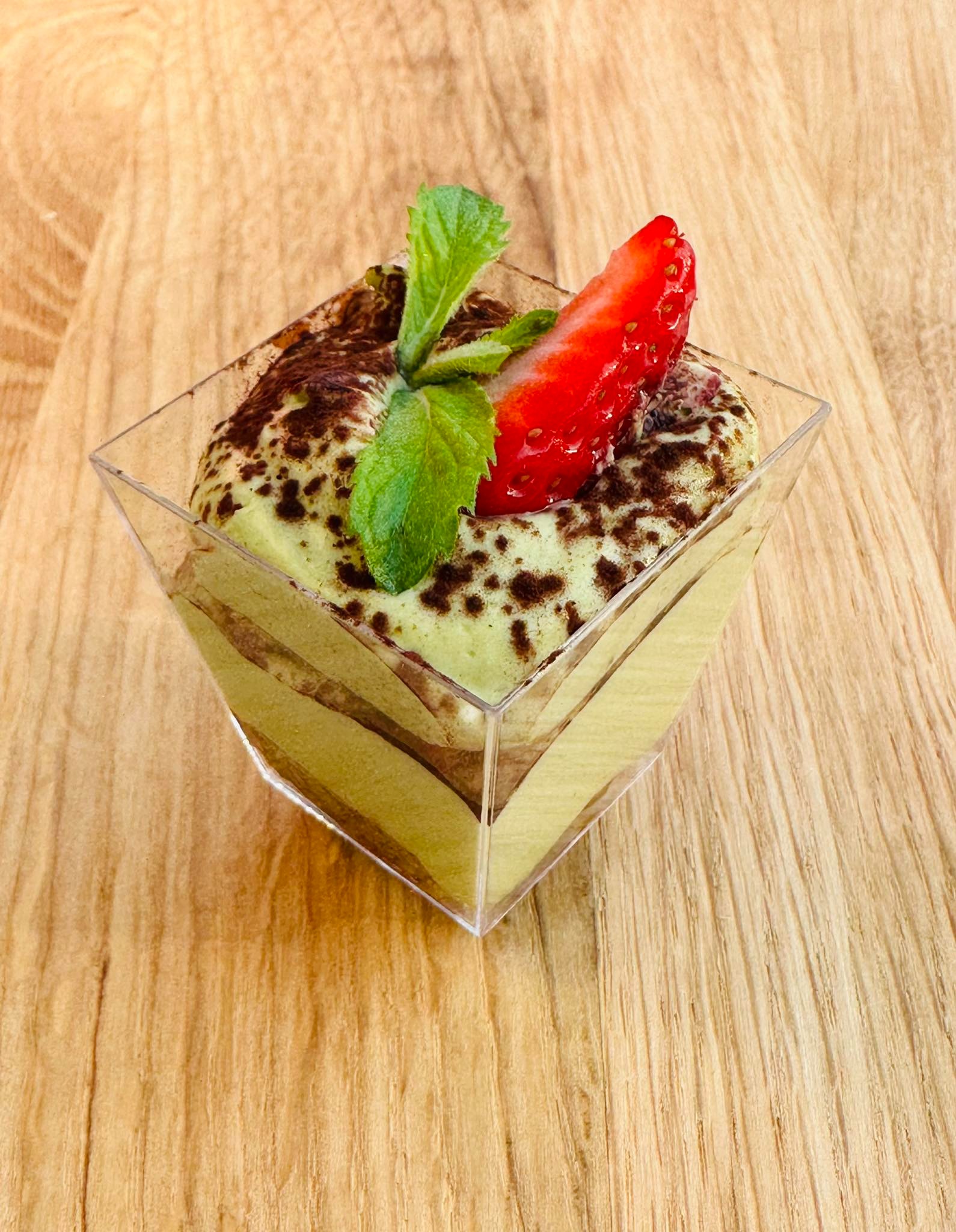 Miniaturowy deser tiramisu o smaku matcha w przezroczystym, kwadratowym naczyniu, udekorowany świeżą truskawką i gałązką mięty, prezentowany na drewnianym blacie.