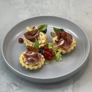 Elegancko podane tartaletki z kremowym nadzieniem, szynką parmeńską, suszonymi pomidorami i świeżymi listkami bazylii, udekorowane czerwoną porzeczką i plasterkiem sera na szarym talerzu.