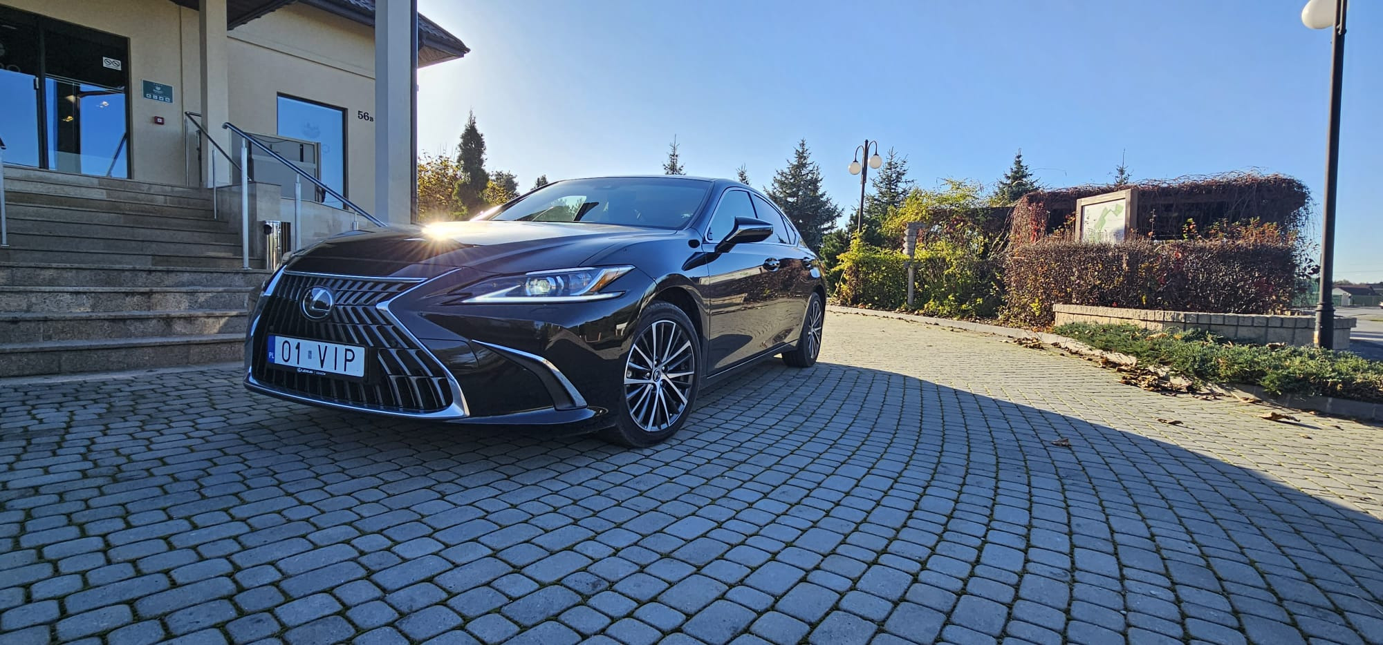 Czarny samochód osobowy Lexus z numerem rejestracyjnym 01 VIP zaparkowany przed budynkiem z schodami i szklanymi drzwiami, na kostce brukowej w słoneczny dzień.