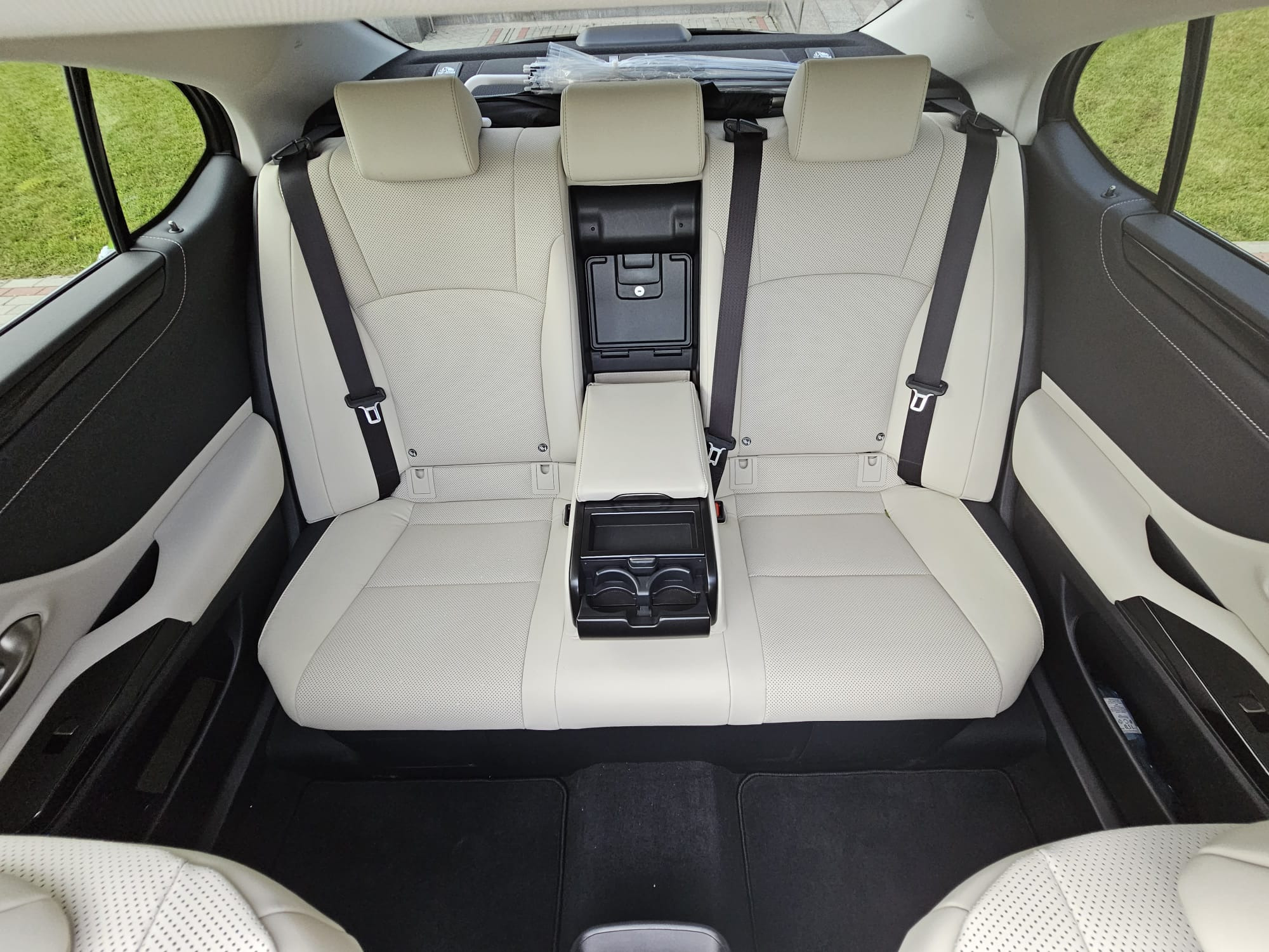 Wnętrze samochodu z jasną, perforowaną skórzaną tapicerką tylnych siedzeń, czarnymi pasami bezpieczeństwa i podłogą, widok z góry na podłokietnik z uchwytami na napoje.