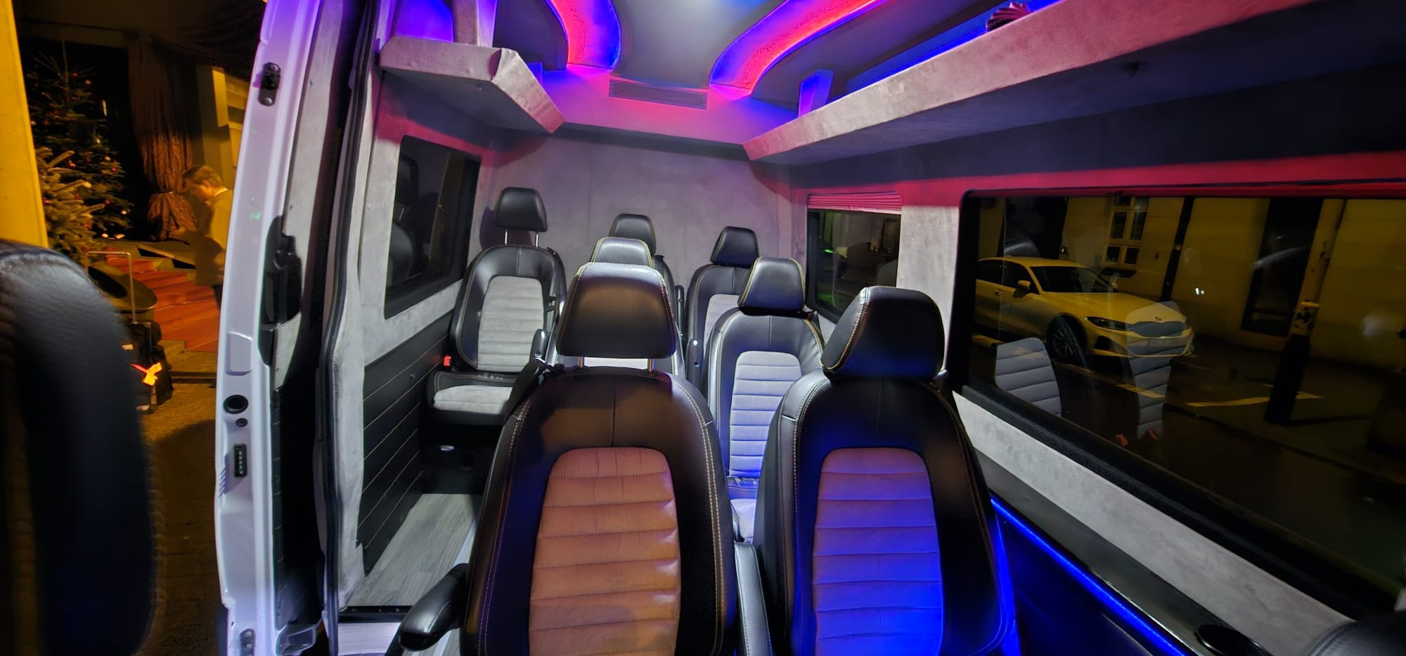 Wnętrze luksusowego busa z podświetleniem LED, skórzane fotele z szarymi wstawkami, widok z otwartych drzwi na oświetlony budynek i zaparkowany samochód.