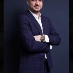 Portret mężczyzny w okularach, granatowym garniturze w kratę i białej koszuli, z założonymi rękami i złotym zegarkiem na tle ciemnoniebieskim.