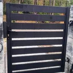 Nowoczesna, pozioma, czarna brama wjazdowa z metalowymi profilami, zamontowana na betonowych słupkach, widok na las w tle.