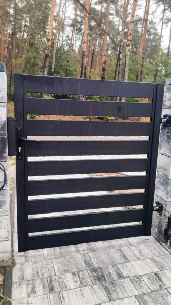 Nowoczesna, pozioma, czarna brama wjazdowa z metalowymi profilami, zamontowana na betonowych słupkach, widok na las w tle.