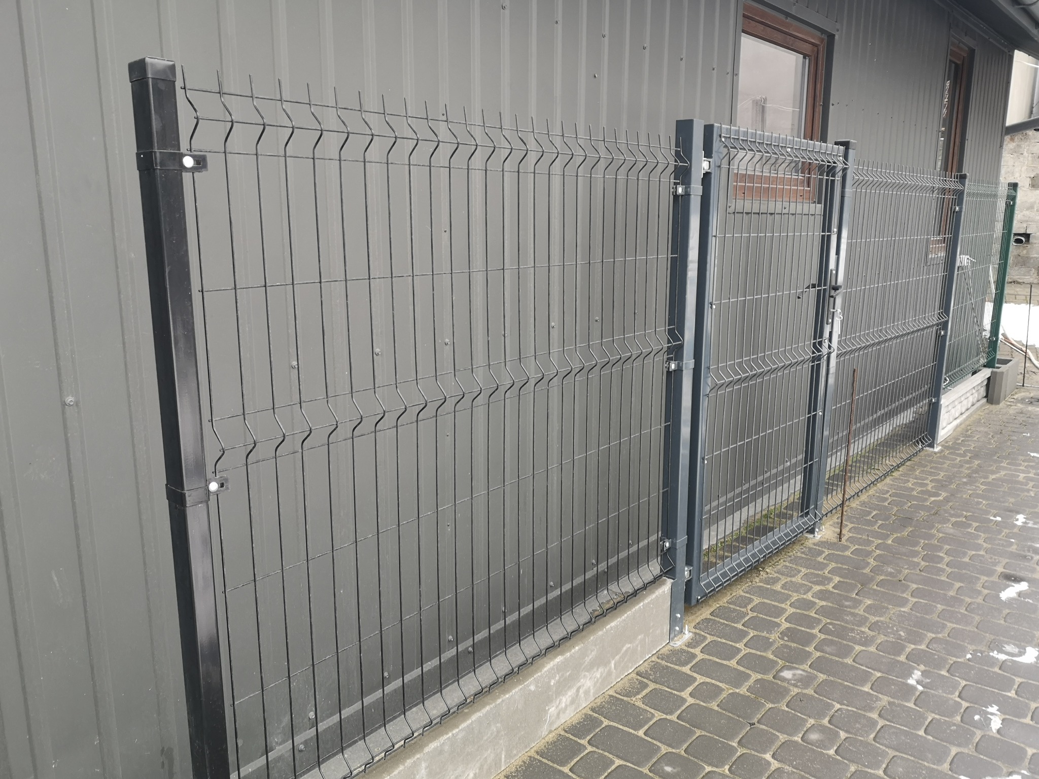 Nowoczesne ogrodzenie panelowe z siatki zgrzewanej w kolorze grafitowym, zamontowane na betonowej podmurówce, przylegające do budynku obłożonego blachą trapezową.