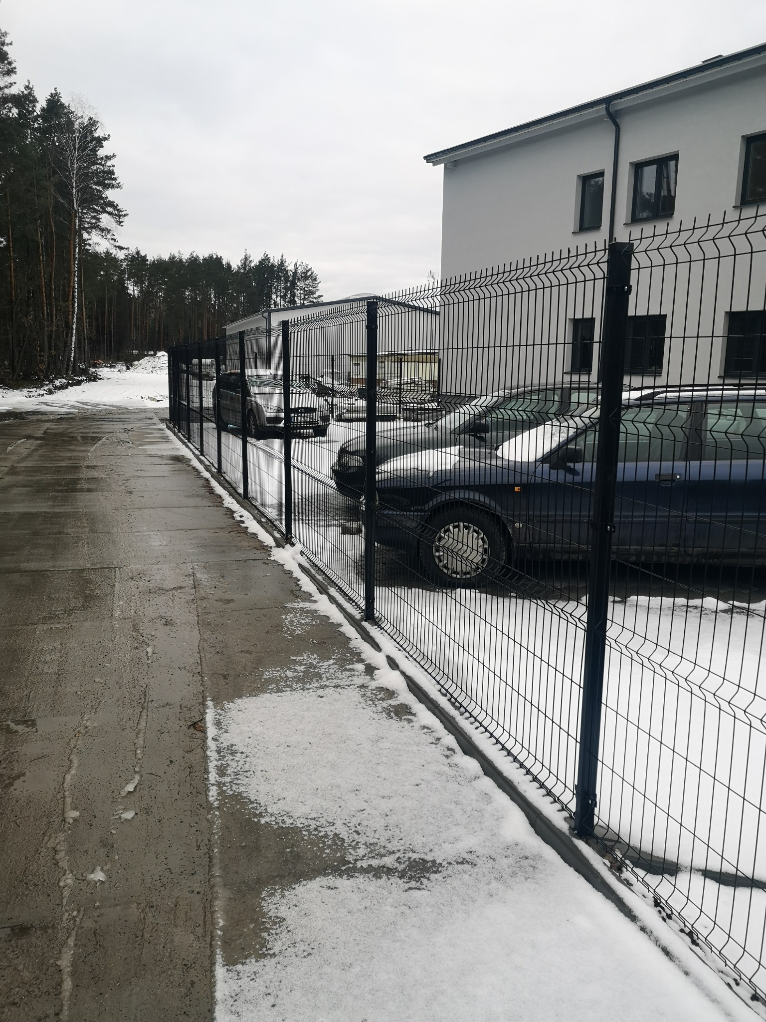 Nowoczesne ogrodzenie panelowe z drutu, oddzielające parking od budynku biurowego w zimowej scenerii, z widocznymi samochodami za ogrodzeniem.