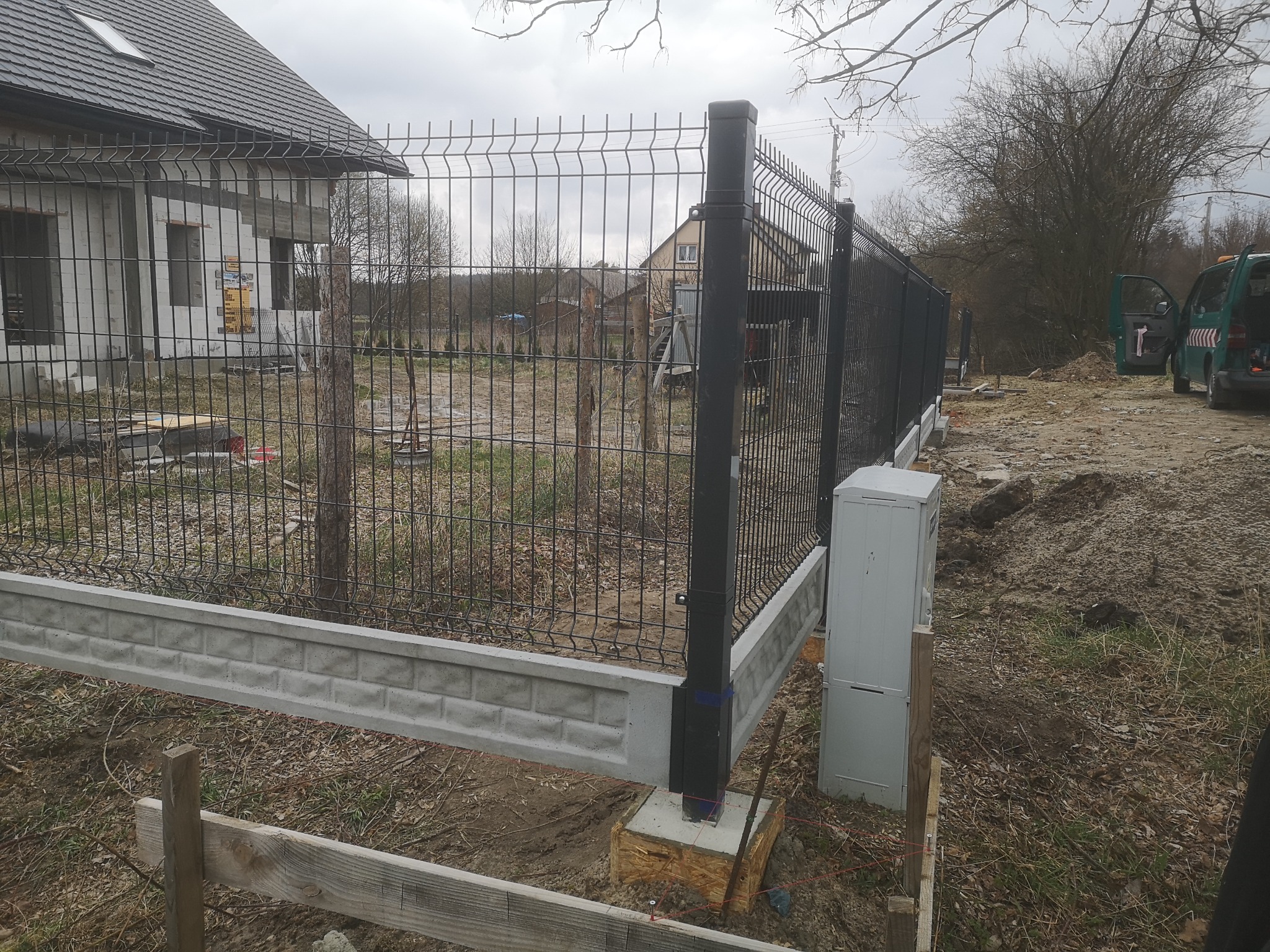 Montaż panelowego ogrodzenia z podmurówką betonową imitującą cegłę, widoczne słupki w kolorze grafitowym i elementy szalunku, w tle dom w trakcie budowy.